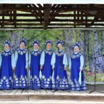 Фотография ансамбля "Заряночка". Девушки в синих народных костюмах, на сцене. Справа стоит гармонист
